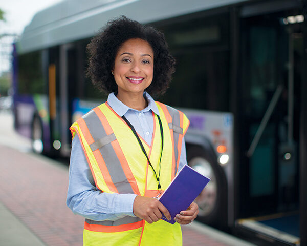 An ambassador wearing a safety vest in front of a parked bus at a transfer station.