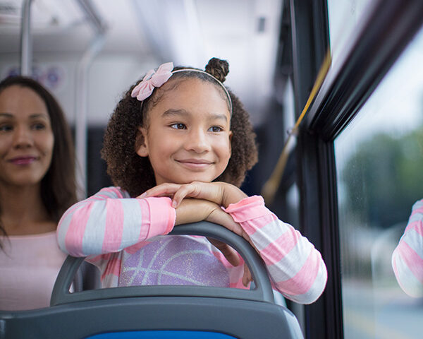 A young girl smiles while looking out of the window on the bus she is riding in.